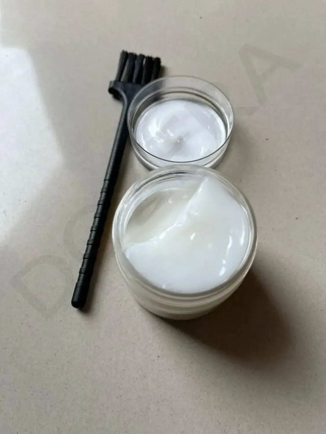 Two containers of white cream with a black brush on a beige surface